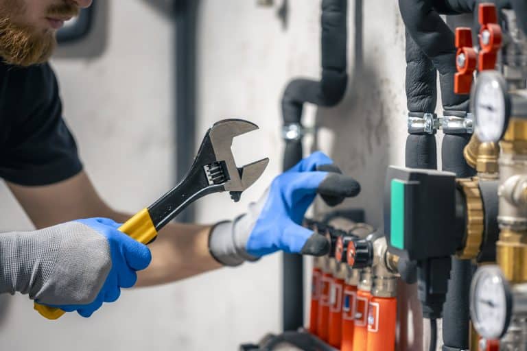 A person wearing blue nitrile gloves uses an adjustable wrench with a yellow and black handle to work on industrial equipment, with gauges, valves, and red components visible in the background of what appears to be a workshop or industrial facility.